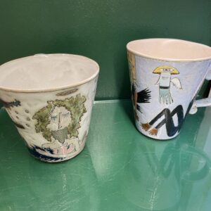 Two "Tass suurem - KAURI KALLAS - K&Otilde;IK" ceramic mugs with hand-painted faces and nature-inspired designs sit on a green reflective surface, capturing the curated feel of a Tallinn concept shop. Gate21.ee