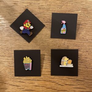 Pins are displayed on black squares on a wooden surface, featuring playful Estonian designs: a red and blue runner, spray cleaner, chips container, and a hedgehog reading&mdash;a quirky touch from the Pins collection. Gate21.ee