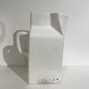 A white ceramic jug shaped like a rectangular milk carton sits on a white surface, with black speckles near its base and a handle on one side&mdash;perfect for adding a curated touch to any concept shop or gift shop display. Gate21.ee