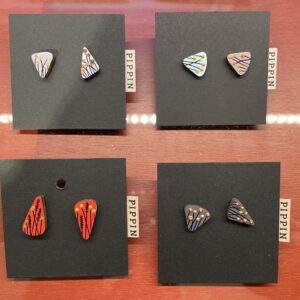 Four pairs of PIPPIN BIRD earrings featuring geometric and botanical designs in red, black, purple, blue, and yellow are displayed on black Pippin cards atop reddish wood&mdash;a curated Estonian design selection ideal for gift shops. Gate21.ee