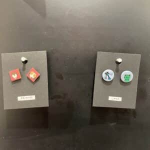 Two pairs of stud earrings by KAURI KALLAS are displayed on black cards: the left, labelled FRIENDS, features red animal-face studs; the right, labelled LAKE, showcases blue and green studs. Product name: Tass nagu + others. Gate21.ee