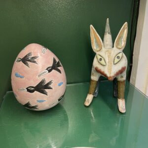 A large pink egg-shaped object decorated with black birds and blue clouds sits next to a ceramic animal figurine, both displayed on a glass shelf in a Tallinn gift shop against a green background. Gate21.ee