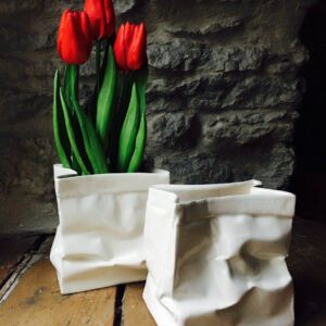Two white paper vases, inspired by Estonian design and shaped like crumpled bags, sit on a wooden surface. One holds three red tulips with green leaves; the other is empty. A stone wall completes this stylish display. Gate21.ee
