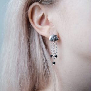 Close-up of a person wearing Artenuf Earrings - K&Otilde;IK: silver cloud-shaped design with three dangling chains tipped with small black beads&mdash;a stylish choice to elevate your look or gift from your favourite concept store. Gate21.ee