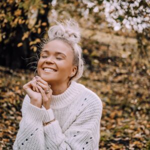 A person with fair hair in a messy bun, wearing a white knitted jumper, sits outdoors on fallen autumn leaves, eyes closed and smiling with hands clasped under chin. A mobile rests on their knee—perhaps after shopping in Tallinn or visiting a concept shop. Gate21.ee