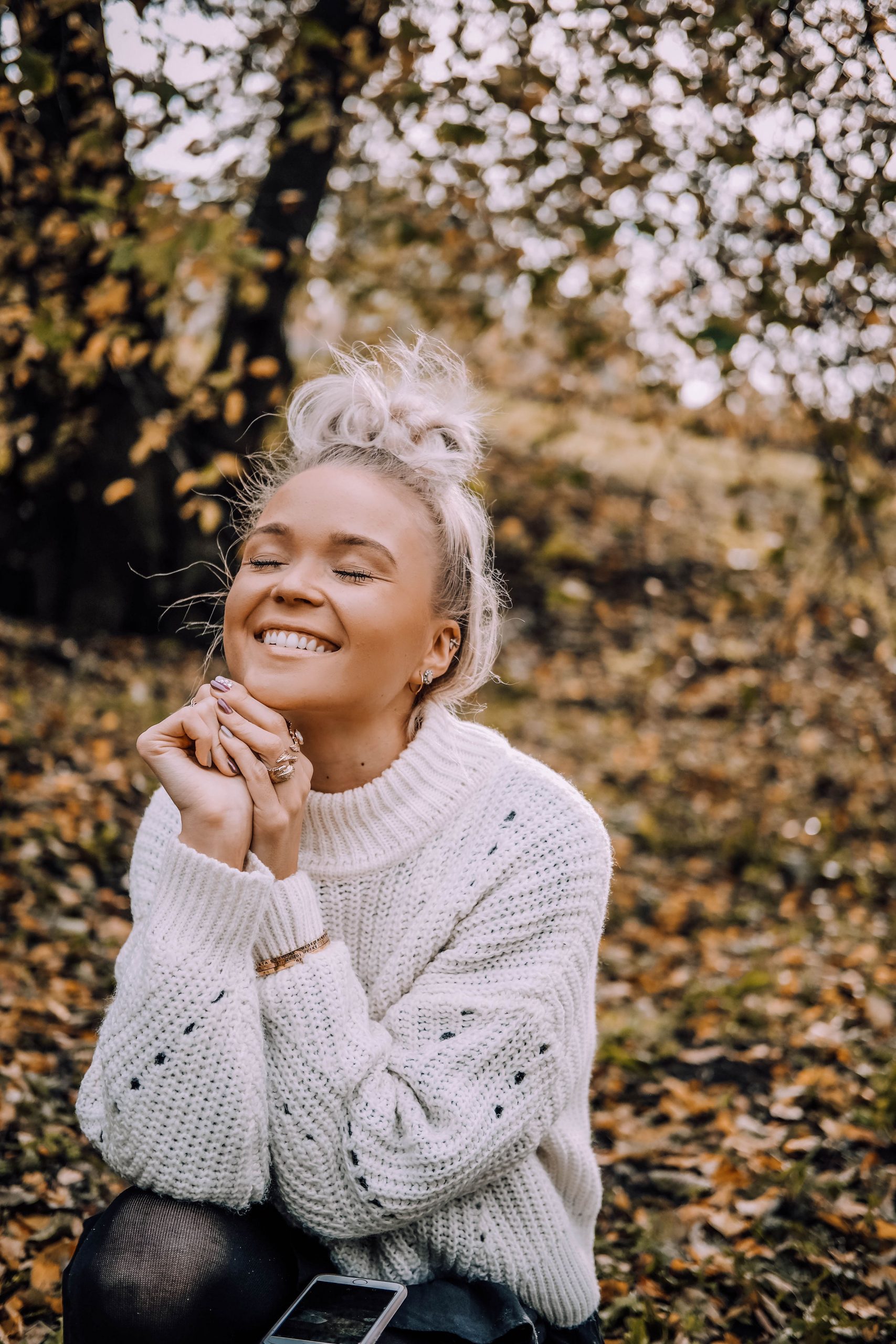 A person with fair hair in a messy bun, wearing a white knitted jumper, sits outdoors on fallen autumn leaves, eyes closed and smiling with hands clasped under chin. A mobile rests on their knee—perhaps after shopping in Tallinn or visiting a concept shop. Gate21.ee