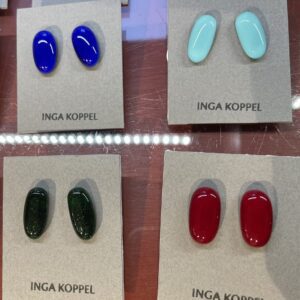 Four pairs of oval glass stud earrings by INGA KOPPEL&mdash;blue, light blue, dark green, and red&mdash;showcase Estonian design. Displayed on grey cards against a reflective surface, they're an ideal Tallinn shopping or concept shop find. Gate21.ee