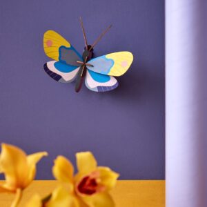 The Yellow Monarch Butterfly, featuring vibrant yellow, blue, and purple wings, is displayed on a purple wall with yellow flowers and a cylindrical purple object—ideal for Tallinn shopping or as a unique gift shop find. Gate21.ee