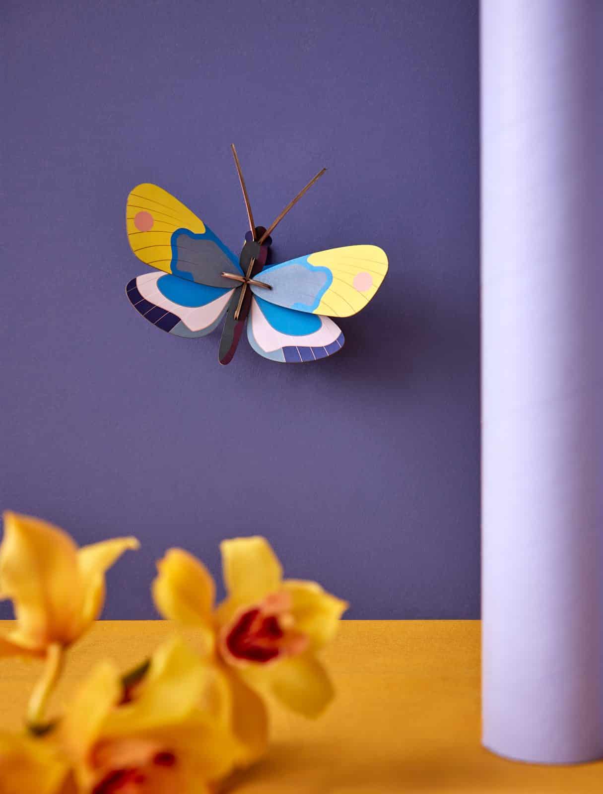 The Yellow Monarch Butterfly, featuring vibrant yellow, blue, and purple wings, is displayed on a purple wall with yellow flowers and a cylindrical purple object—ideal for Tallinn shopping or as a unique gift shop find. Gate21.ee