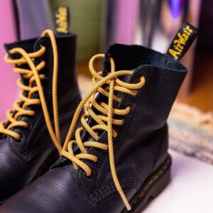 A close-up of black Dr. Martens boots with yellow stitching and Reflective Rusty Laces by Extrawize, shown on a light surface with a blurred background—a striking look that showcases innovative footwear style. Gate21.ee