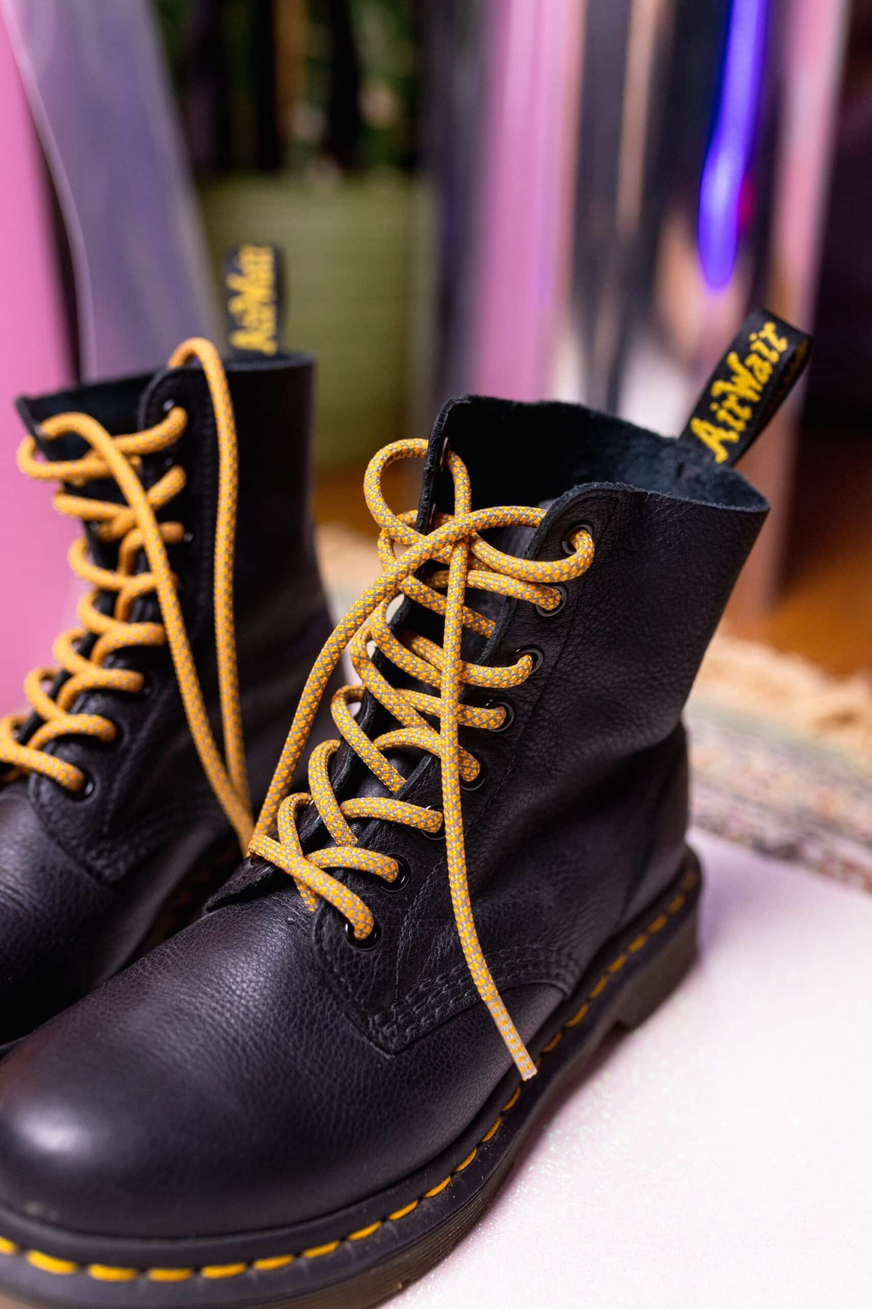 A close-up of black Dr. Martens boots with yellow stitching and Reflective Rusty Laces by Extrawize, shown on a light surface with a blurred background—a striking look that showcases innovative footwear style. Gate21.ee