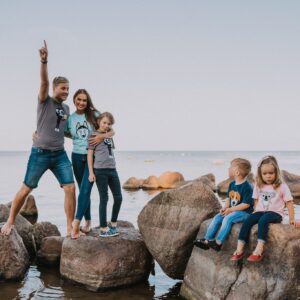 A man, woman, and three children stand and sit on large rocks by the water. Dressed in curated shirts with cartoon animal prints, they gaze in different directions under a clear sky, embodying the unique ideas of a concept store family photoshoot. Gate21.ee