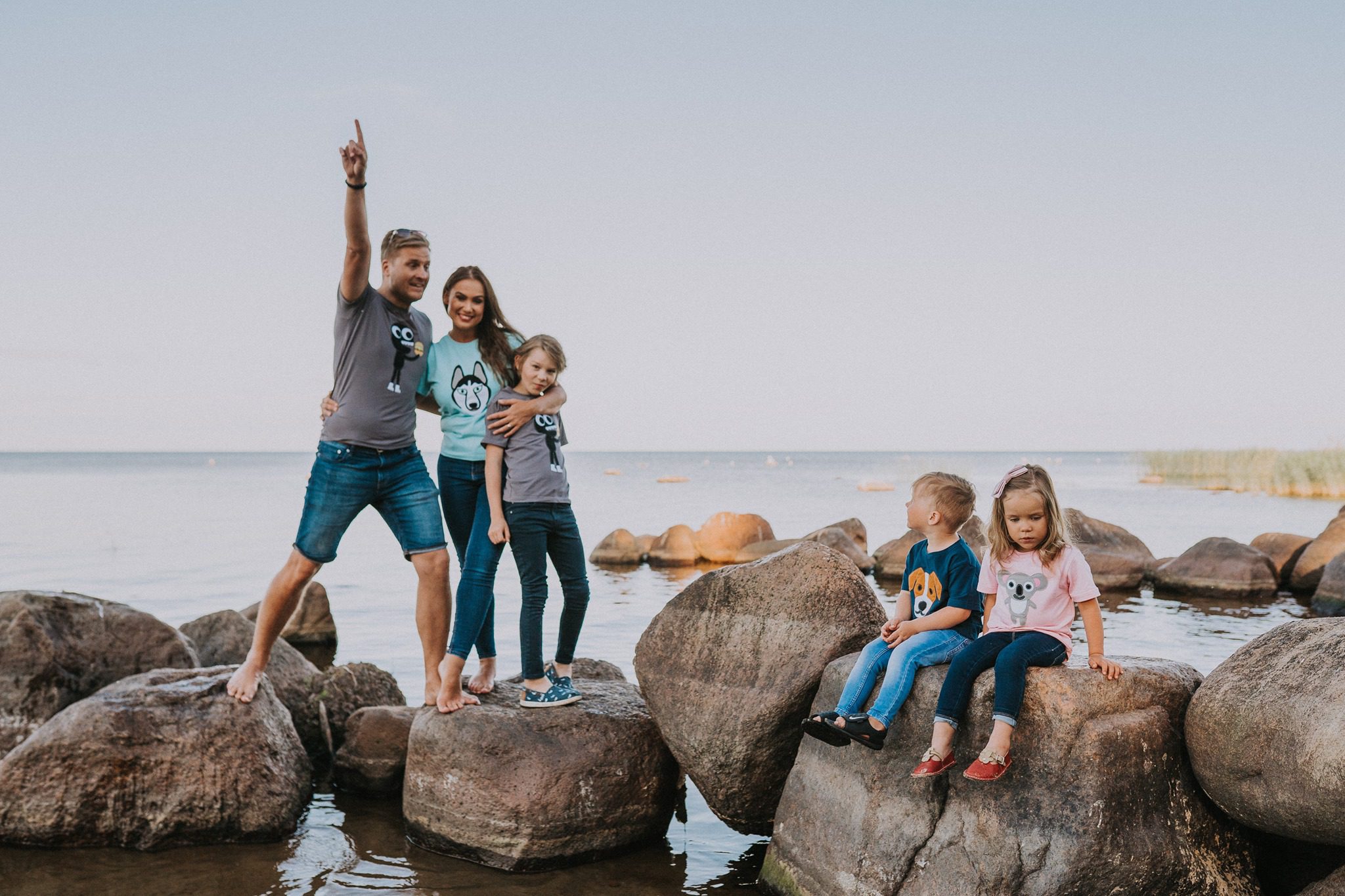 A man, woman, and three children stand and sit on large rocks by the water. Dressed in curated shirts with cartoon animal prints, they gaze in different directions under a clear sky, embodying the unique ideas of a concept store family photoshoot. Gate21.ee
