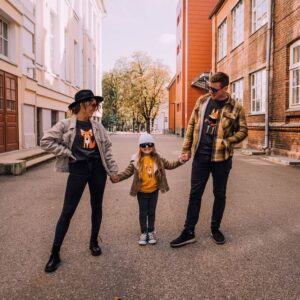 Three people—two adults and a child—stand on a paved street holding hands. All wear matching blue Guitar sweatshirts, jackets, and sunglasses as the adults look fondly at the child between them. Gate21.ee