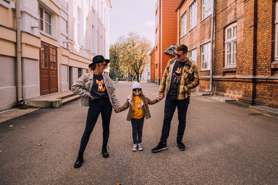 Three people—two adults and a child—stand on a paved street holding hands. All wear matching blue Guitar sweatshirts, jackets, and sunglasses as the adults look fondly at the child between them. Gate21.ee
