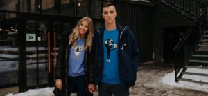 A woman and a man stand outside a concept shop holding hands, both wearing Dino - Sweatshirt Black with cartoon designs. Snow covers the ground near a staircase and a building with glass doors. Gate21.ee