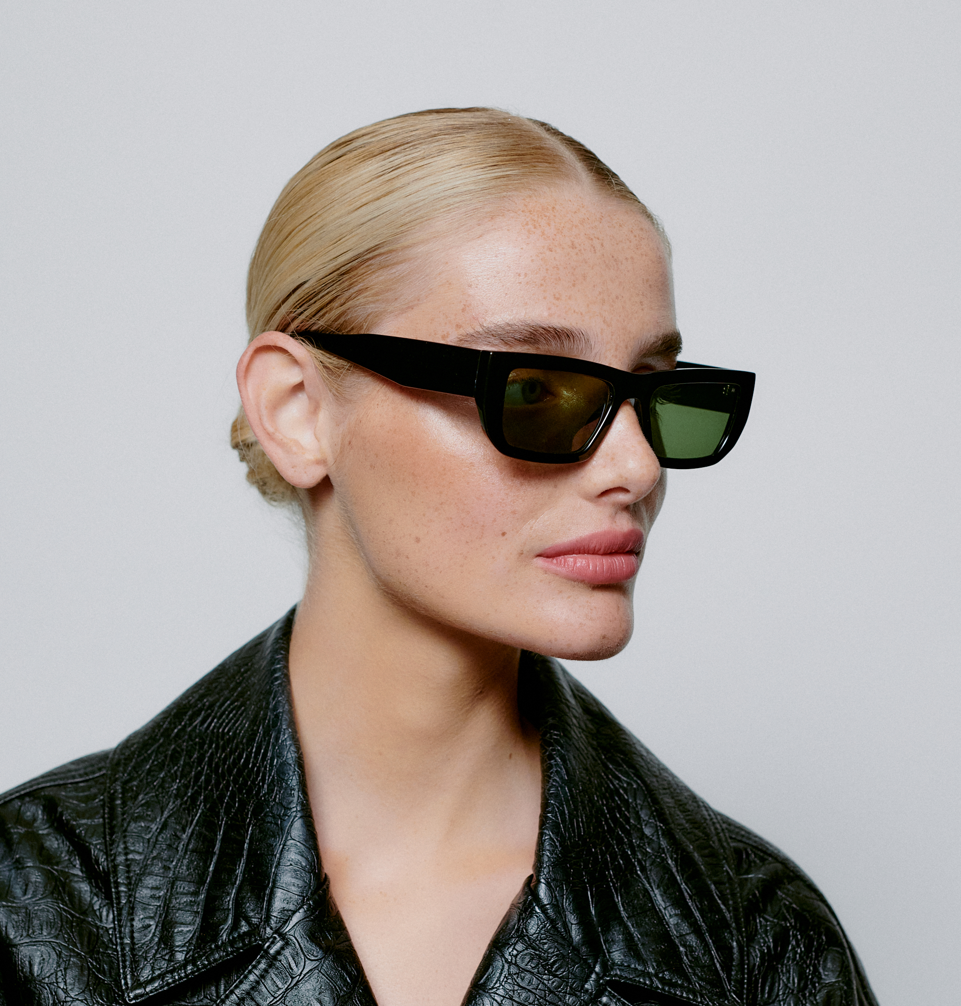 A person with blonde hair pulled back wears FAME - Black rectangular sunglasses and a black textured jacket, facing slightly left against a plain grey background—embracing the understated style of Tallinn's curated concept stores. Gate21.ee