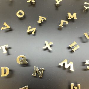 Scattered gold metal letters from LETTERS by F - K&Otilde;IK rest on a smooth, dark surface, reflecting Estonian design. Arranged in various orientations, these letters form no specific words or patterns. Gate21.ee
