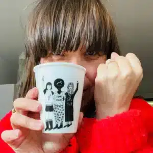 A brown-haired person with fringe in a bright red jumper holds the 'Equal Rights' paper cup—white, featuring illustrations of women and the words GRL PWR and GOAL DIGGER—ideal for discovering unique Estonian design at a concept shop. Gate21.ee