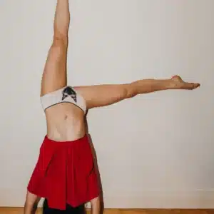 A person does a headstand with arms and legs extended, wearing a red skirt and Siamese Cat Knickers featuring a bow, set against a plain white wall and wooden floor—a scene that highlights distinctive style. Gate21.ee