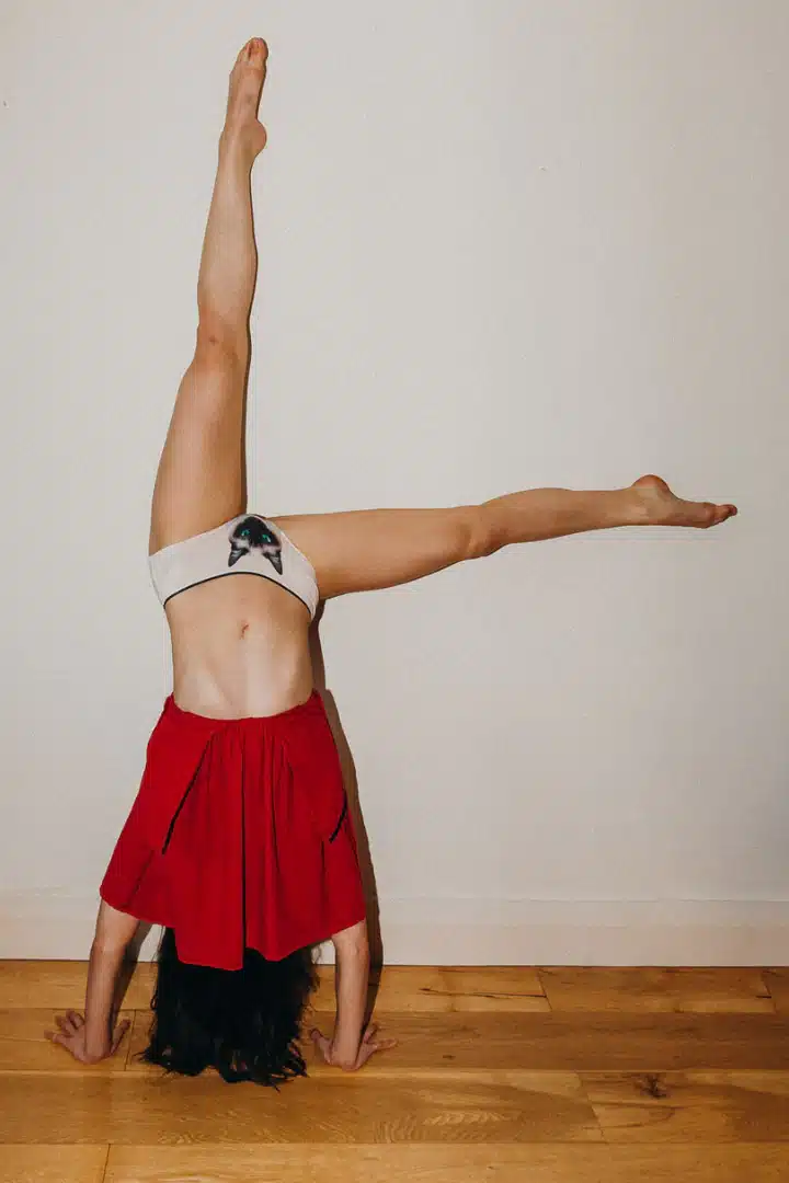 A person does a headstand with arms and legs extended, wearing a red skirt and Siamese Cat Knickers featuring a bow, set against a plain white wall and wooden floor—a scene that highlights distinctive style. Gate21.ee