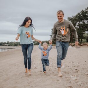 A woman, a man, and a young child wear matching DJ - T-shirt Red shirts from a unique gift shop as they walk barefoot, holding hands on a sandy beach with trees and cloudy skies in the background. Gate21.ee