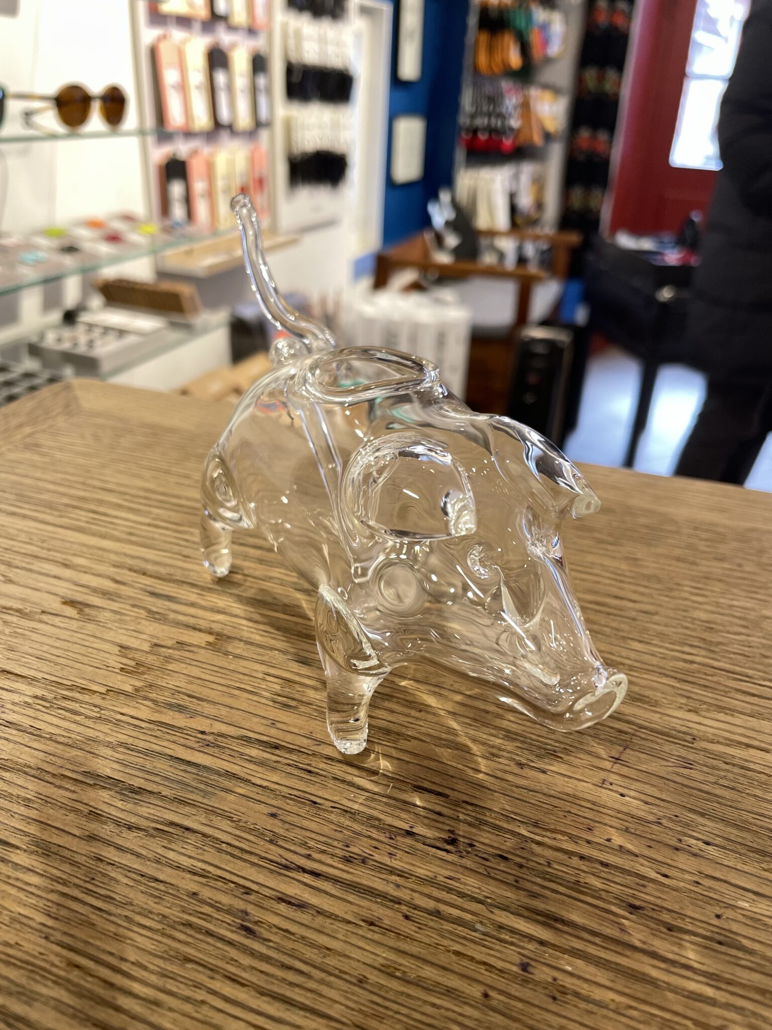 The Konjakipiip - LOOM, a clear glass pig figurine, rests on a wooden surface in a curated gift shop, surrounded by shelves of Estonian design items. Gate21.ee
