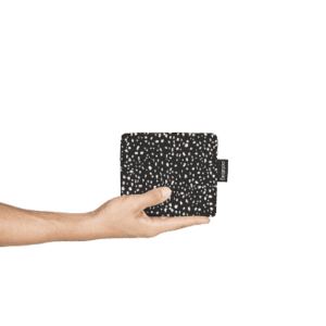 A hand holds the Black Brush—a black rectangular pouch with a white speckled pattern and a side tag—against a white background. This elegant piece of Estonian design is ideal for anyone seeking unique gifts. Gate21.ee