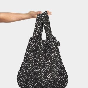 A hand holds up the Black Brush reusable shopping bag, featuring white speckled dots and modern Estonian design, against a plain light grey background—ideal for any concept store. Gate21.ee
