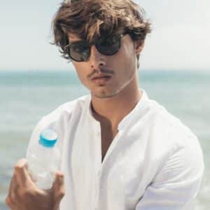 A man in a white shirt stands by the sea, holding a plastic water bottle and wearing "Isla" Black Sunglasses. The calm sea and clear sky behind him embody the curated simplicity of Estonian design. Gate21.ee