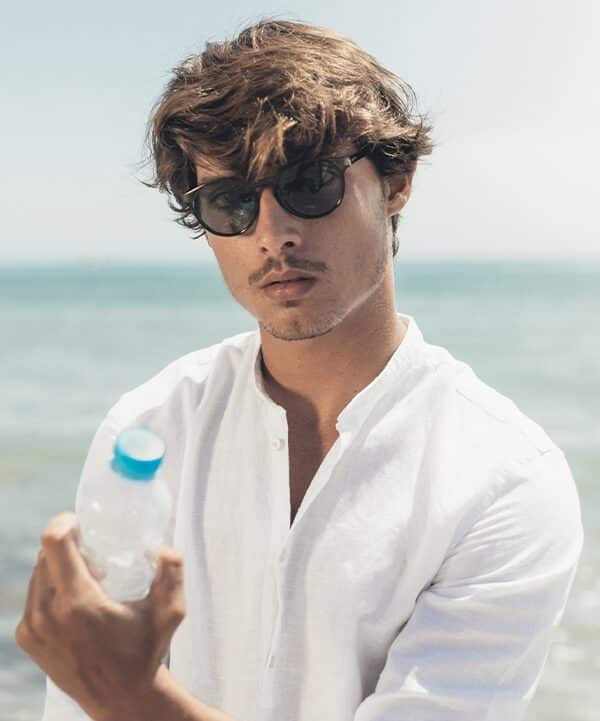 A man in a white shirt stands by the sea, holding a plastic water bottle and wearing "Isla" Black Sunglasses. The calm sea and clear sky behind him embody the curated simplicity of Estonian design. Gate21.ee