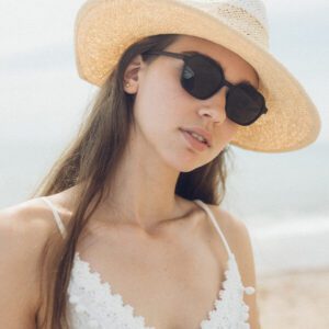 A woman stands outdoors, possibly at the seaside, in a wide-brimmed straw hat and white lace top, wearing "Valle" Black Sunglasses—her look inspired by unique Estonian design, with sand and sky softly blurred in the background. Gate21.ee