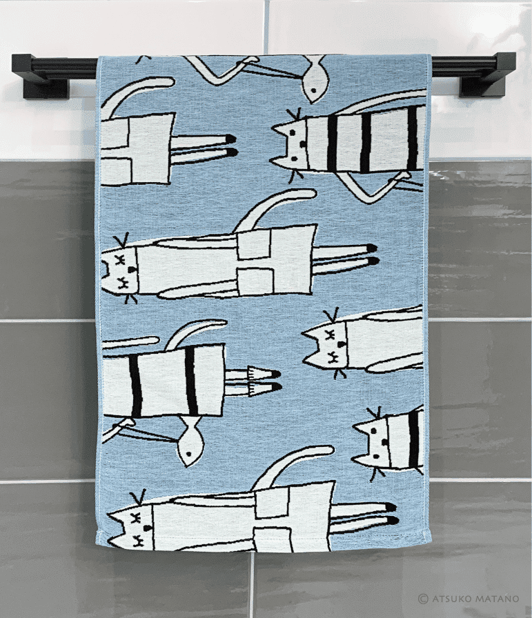 The Cat in New York - KÕIK hand towel, featuring a blue design with white cartoon cats and black outlines, hangs on a black rack against grey tiles—ideal for adding playful charm to your gift shop or home. Gate21.ee