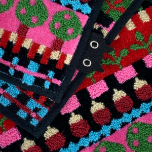 A close-up of colourful, patterned fabric featuring trees and mushrooms. The corner is folded, revealing a black cat face with round white eyes peeking out—a playful touch of curated Estonian design perfect for any gift shop. Gate21.ee