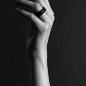 A greyscale photo features a raised hand with slender fingers adorned with the DUALITY RING - BIG "SEOUL," a rectangular black piece that adds an elegant touch of Estonian design against a plain, dark background. Gate21.ee