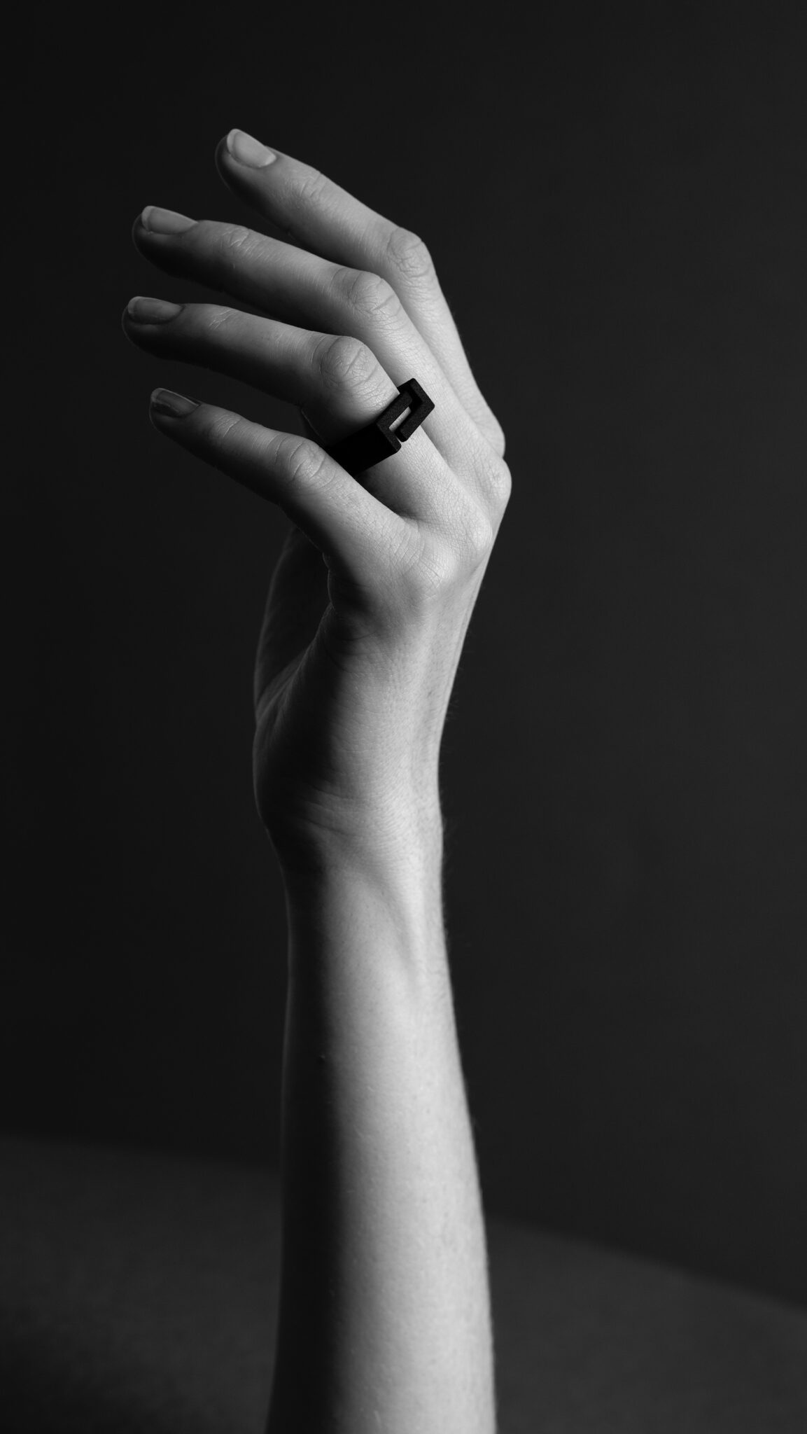 A greyscale photo features a raised hand with slender fingers adorned with the DUALITY RING - BIG "SEOUL," a rectangular black piece that adds an elegant touch of Estonian design against a plain, dark background. Gate21.ee