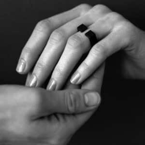 A person gently holds another's hand, which is adorned with the DUALITY RING - JC—an elegant black geometric piece. The black and white photo highlights timeless style reminiscent of a curated concept shop. Gate21.ee