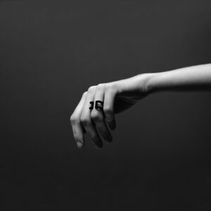 A person with slender fingers extends their hand against a dark background, wearing the bold, angular DUALITY RING - DAN on the middle finger. The high-contrast lighting highlights the ring’s minimalist Estonian design. Gate21.ee