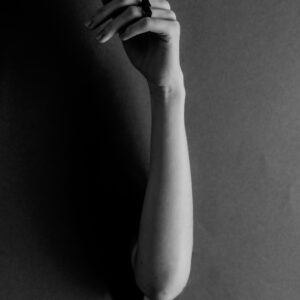 A curated greyscale photo shows a single arm extending through a hole, hand gently bent upward with relaxed fingers. The plain background emphasises the DUALITY RING - HOLL worn on the person's index finger. Gate21.ee