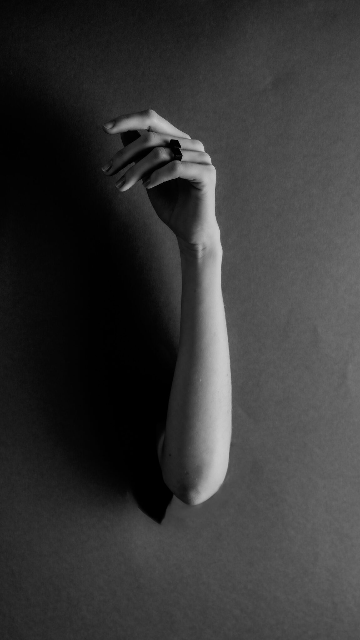A curated greyscale photo shows a single arm extending through a hole, hand gently bent upward with relaxed fingers. The plain background emphasises the DUALITY RING - HOLL worn on the person's index finger. Gate21.ee