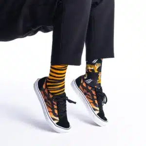A person sits against a white background, wearing black trousers and "Feet of the Tiger" trainers—curated black footwear featuring an orange and brown pattern—paired with mismatched socks: one yellow and black striped, the other with tiger illustrations. Gate21.ee