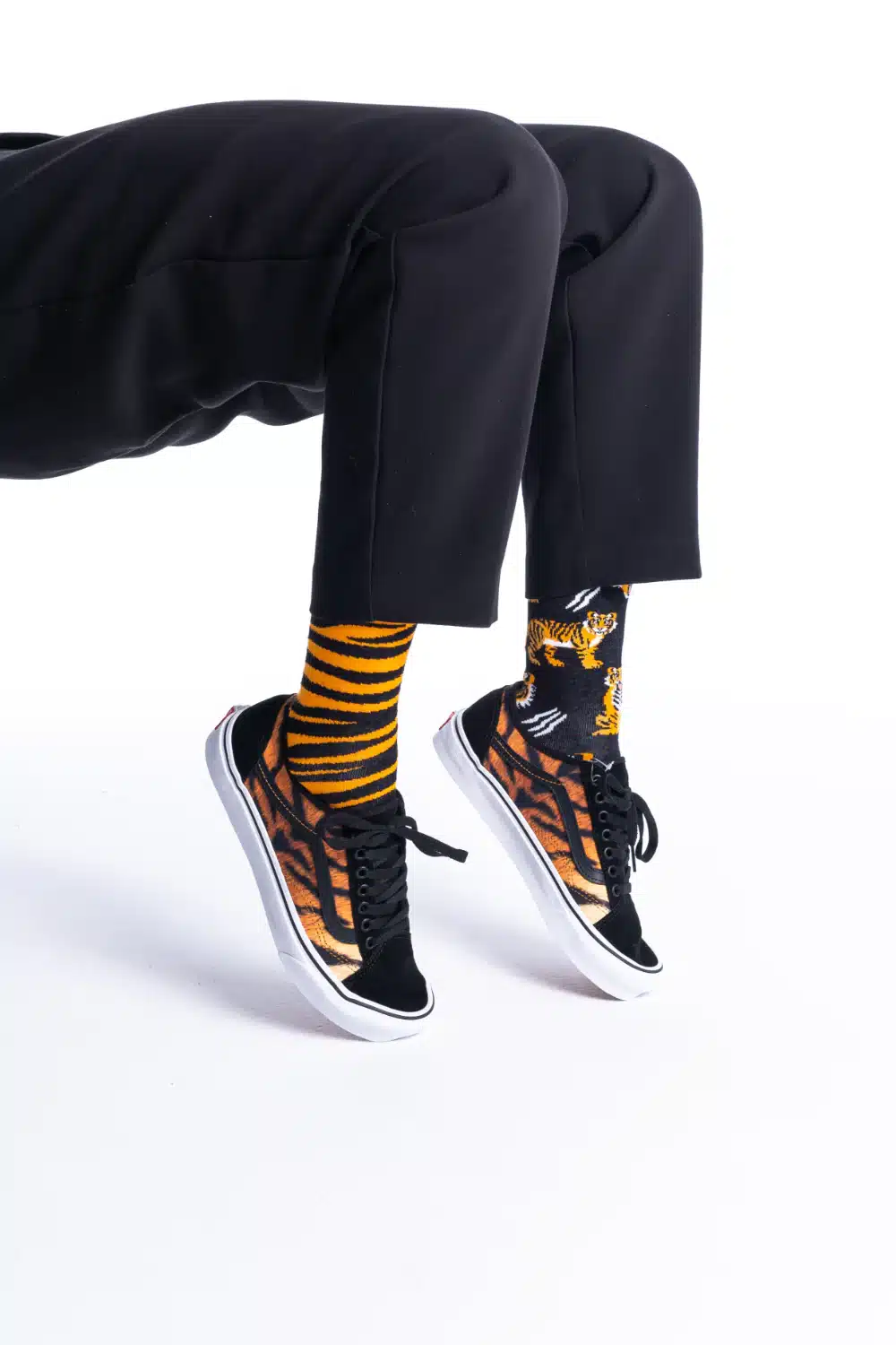 A person sits against a white background, wearing black trousers and "Feet of the Tiger" trainers—curated black footwear featuring an orange and brown pattern—paired with mismatched socks: one yellow and black striped, the other with tiger illustrations. Gate21.ee