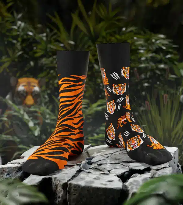 Feet of the Tiger socks—one tiger-striped, one black with playful tiger faces—are showcased on rocks in lush outdoors. A blurred tiger statue adds wild flair, making these socks perfect for unique style or as a standout gift shop feature. Gate21.ee