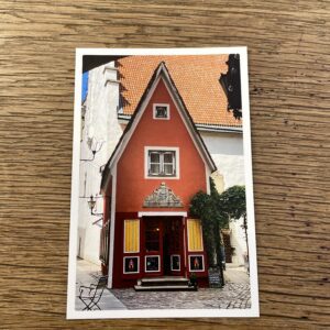 The Red House Photo - Postcard features a red, narrow two-storey building with ornate details and yellow-striped awnings set on cobblestone pavement, evoking the charm of a Tallinn shopping concept store. Gate21.ee