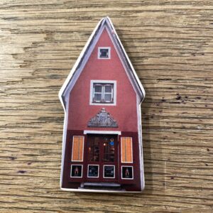 Red House - Magnet: a tall, narrow red house with yellow shutters, ornate door detail, and two windows&mdash;an eye-catching piece of Estonian design to brighten your space. Perfect for adding unique charm to any wooden surface. Gate21.ee