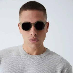 Wearing "Arroyo" Moss Sunglasses and a light grey textured jumper, a short-haired individual stands against a plain background, evoking the chic vibe of a curated Tallinn gift shop whilst looking directly at the camera. Gate21.ee