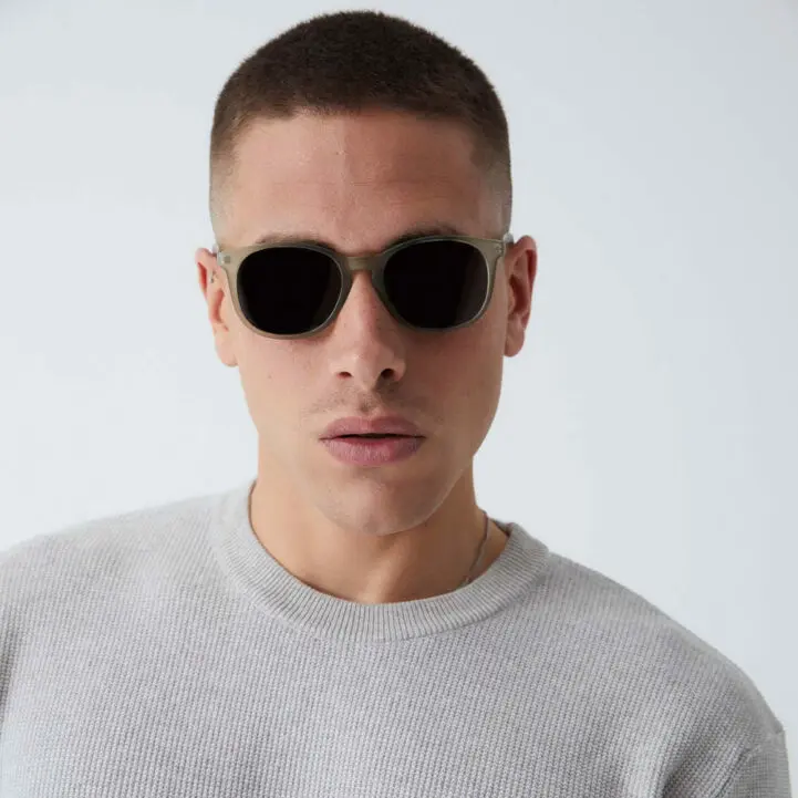 Wearing "Arroyo" Moss Sunglasses and a light grey textured jumper, a short-haired individual stands against a plain background, evoking the chic vibe of a curated Tallinn gift shop whilst looking directly at the camera. Gate21.ee