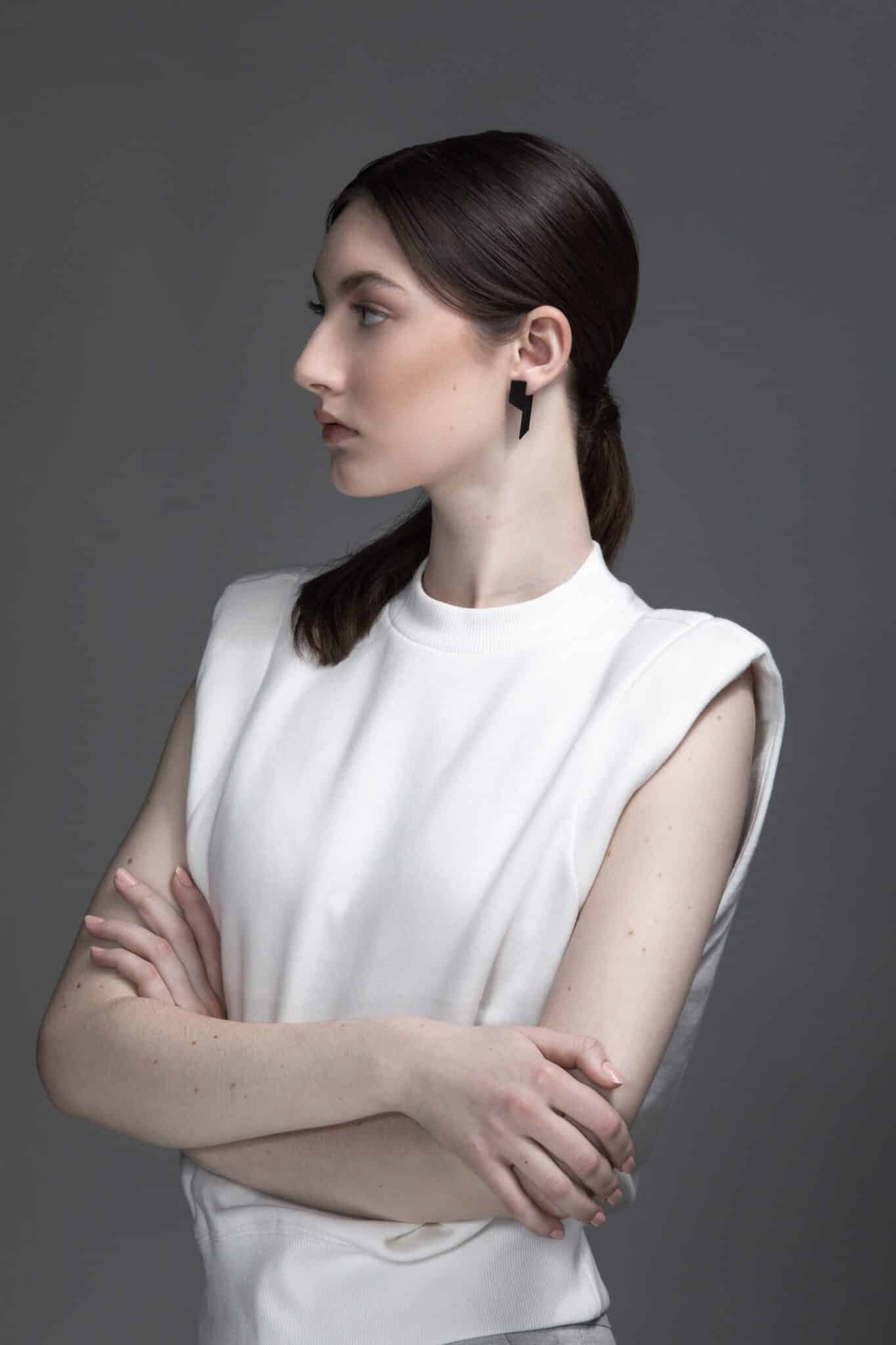 A woman with pale skin and dark brown hair pulled back is standing against a grey background. She wears a sleeveless white top, black geometric earrings—perfect for showcasing Estonian design found in Tallinn shopping concept shops. Gate21.ee