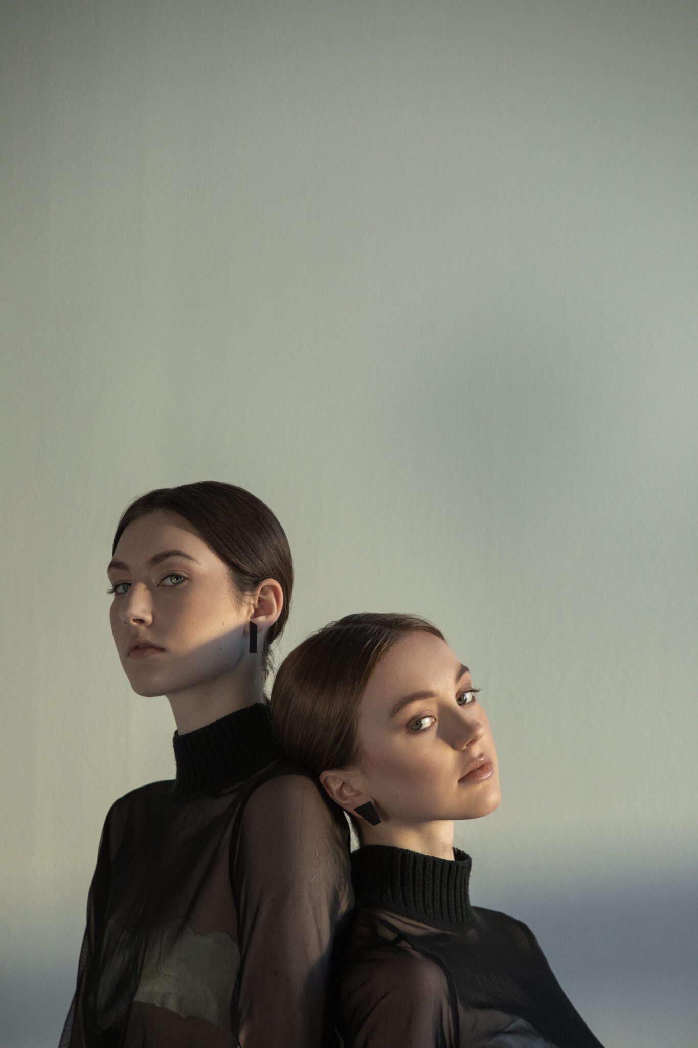 Two women with dark hair in low buns stand together against a plain backdrop, both wearing DUALITY EARRINGS – PARALLEL. Their poised style embodies Estonian elegance, making these earrings ideal for a modern gift shop display. Gate21.ee