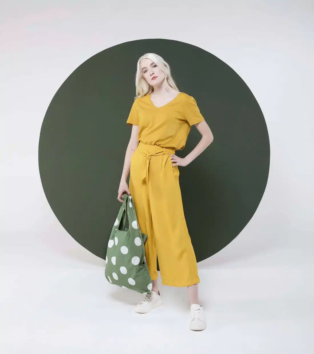 A person with long blonde hair in a mustard yellow outfit and white trainers stands before a large dark green circle, holding the Olive Dots green polka dot bag against a white background. Gate21.ee
